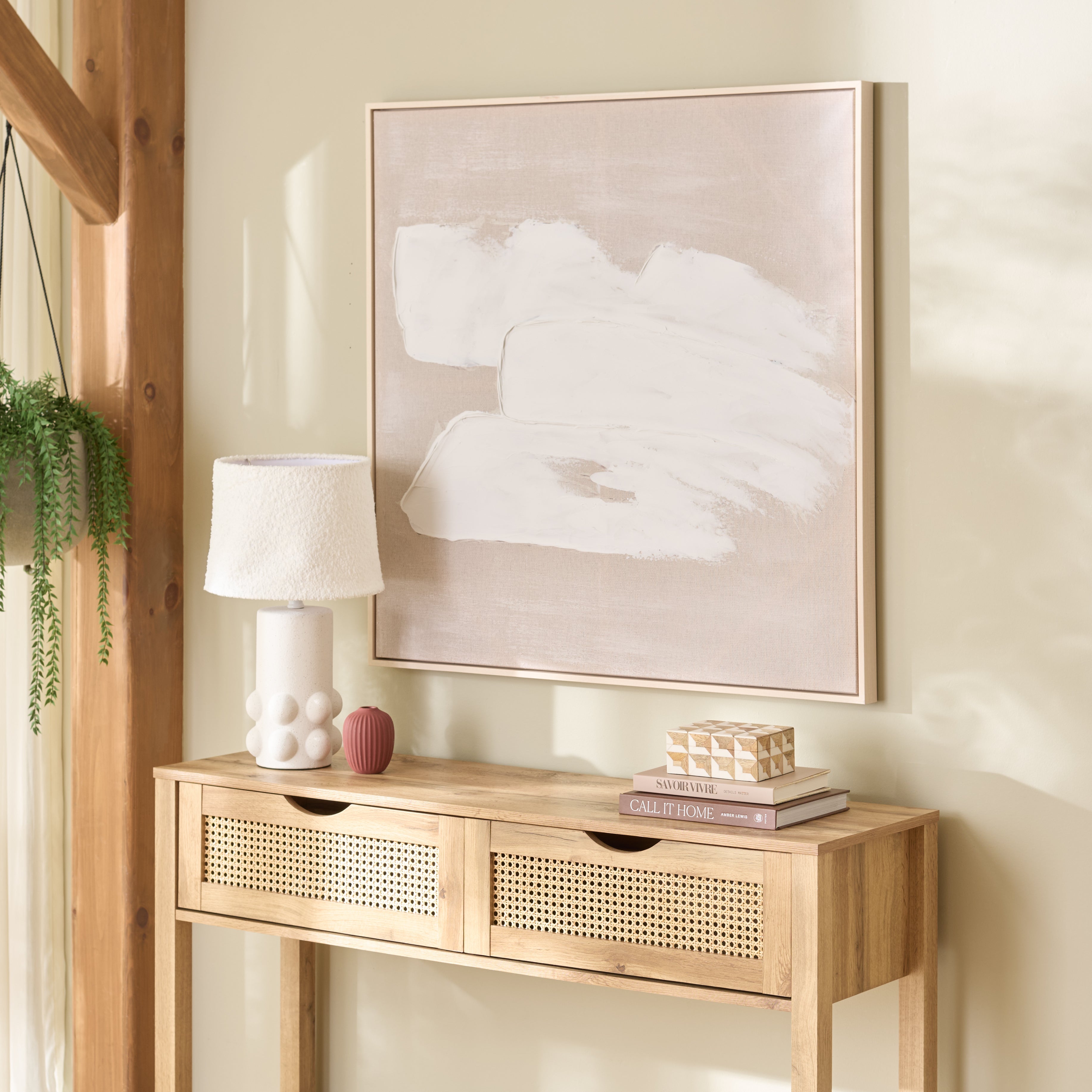 Three Strokes abstract framed wall art styled above a rattan console table in a warm, natural-light entryway vignette with boucle lamp, ceramic vase, books, and trailing plant — Rachel Blindauer