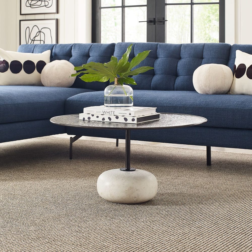 Round coffee table with hammered steel top and carved white stone base, styled in a Mid Century modern living room with layered textures and navy sofa.
