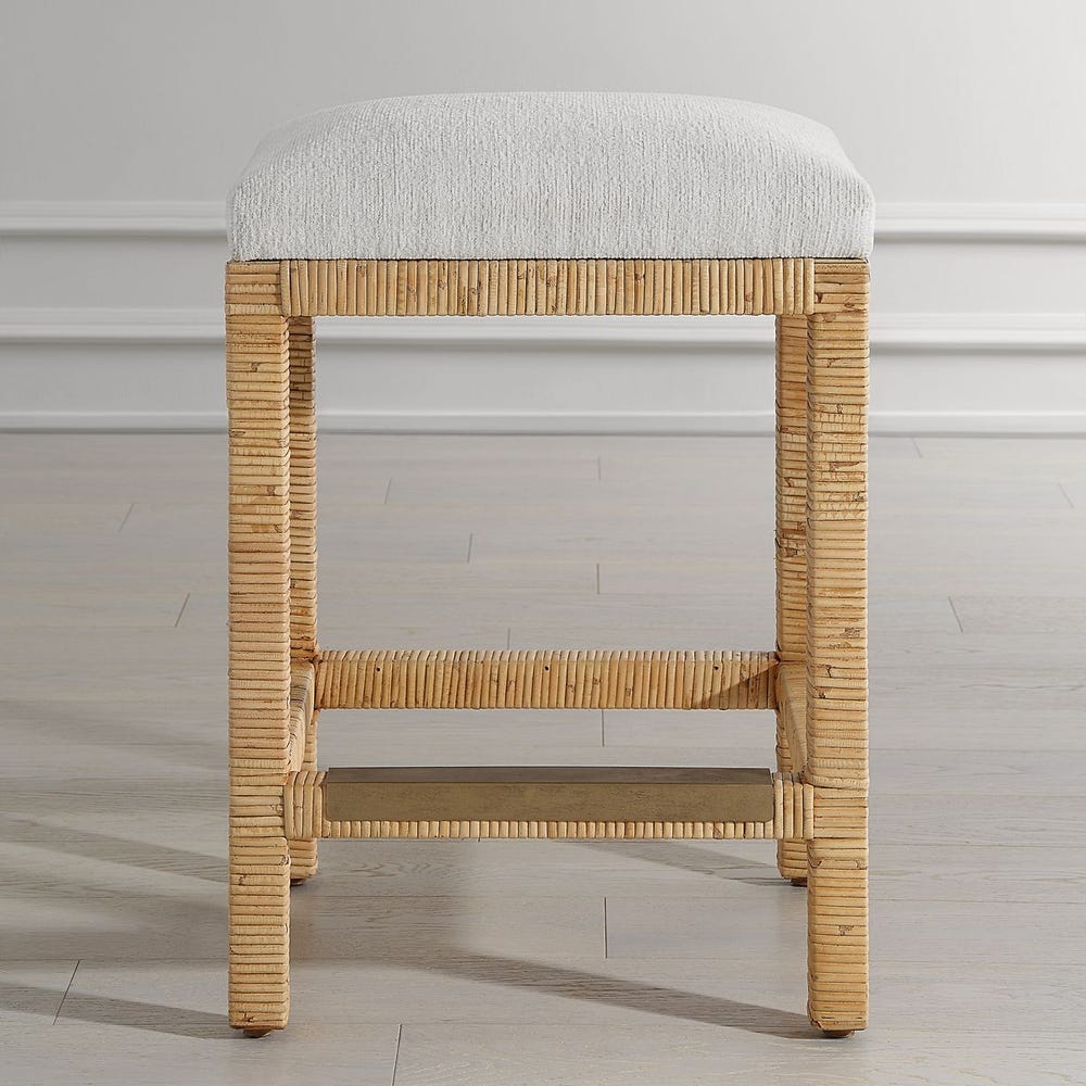Handwoven rattan counter-height stool with off-white cushion and antique gold kickplate—coastal-inspired and refined.