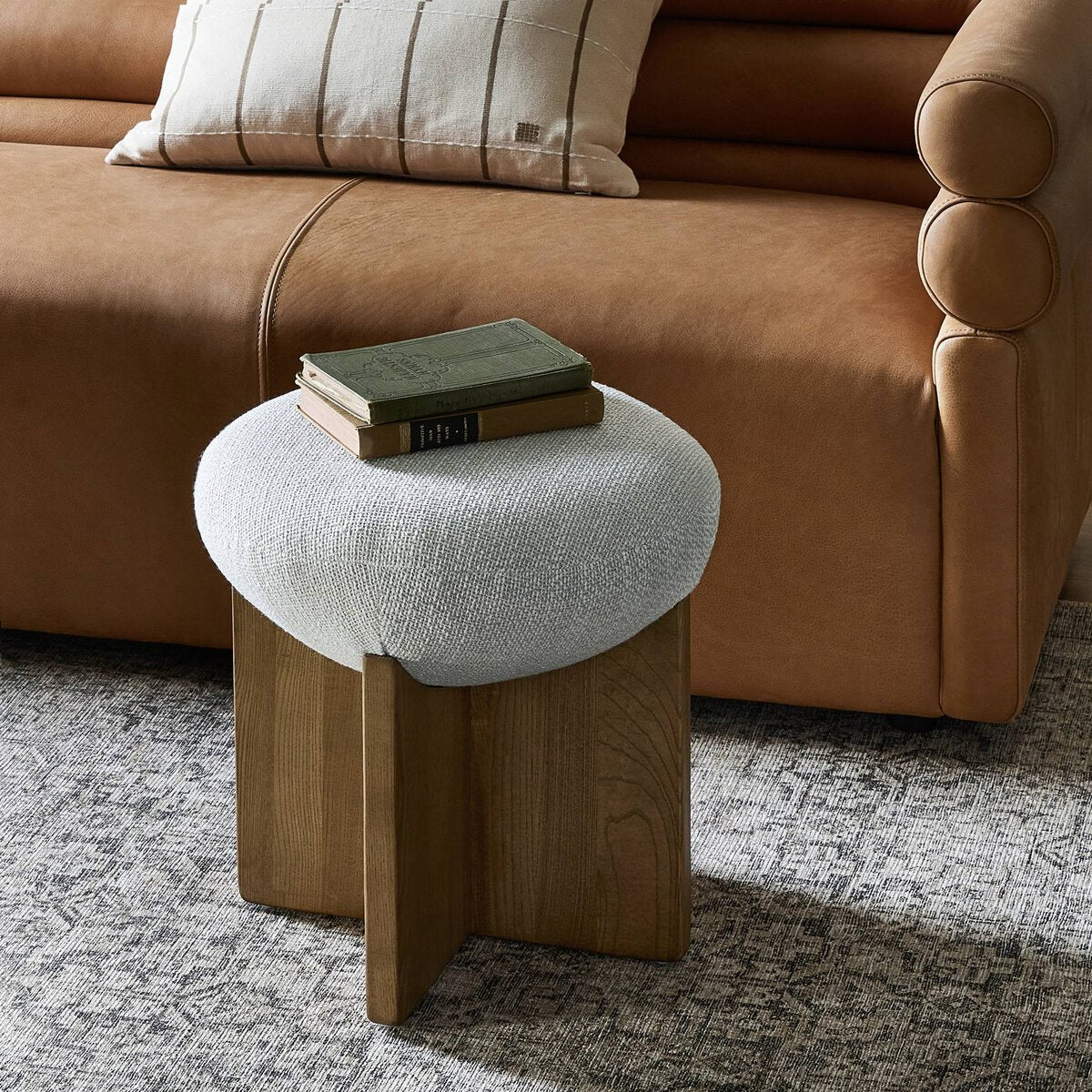 Small round foot stool with a textured white cushion on a wooden base in front of a brown sofa.