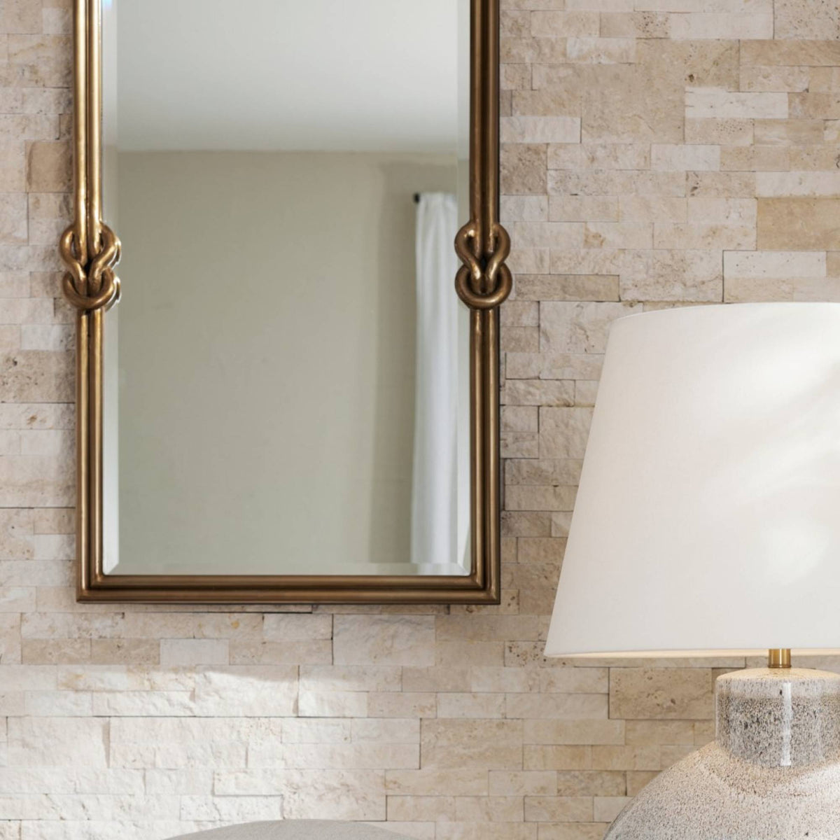 Brass knot mirror hung on a travertine wall above a table lamp