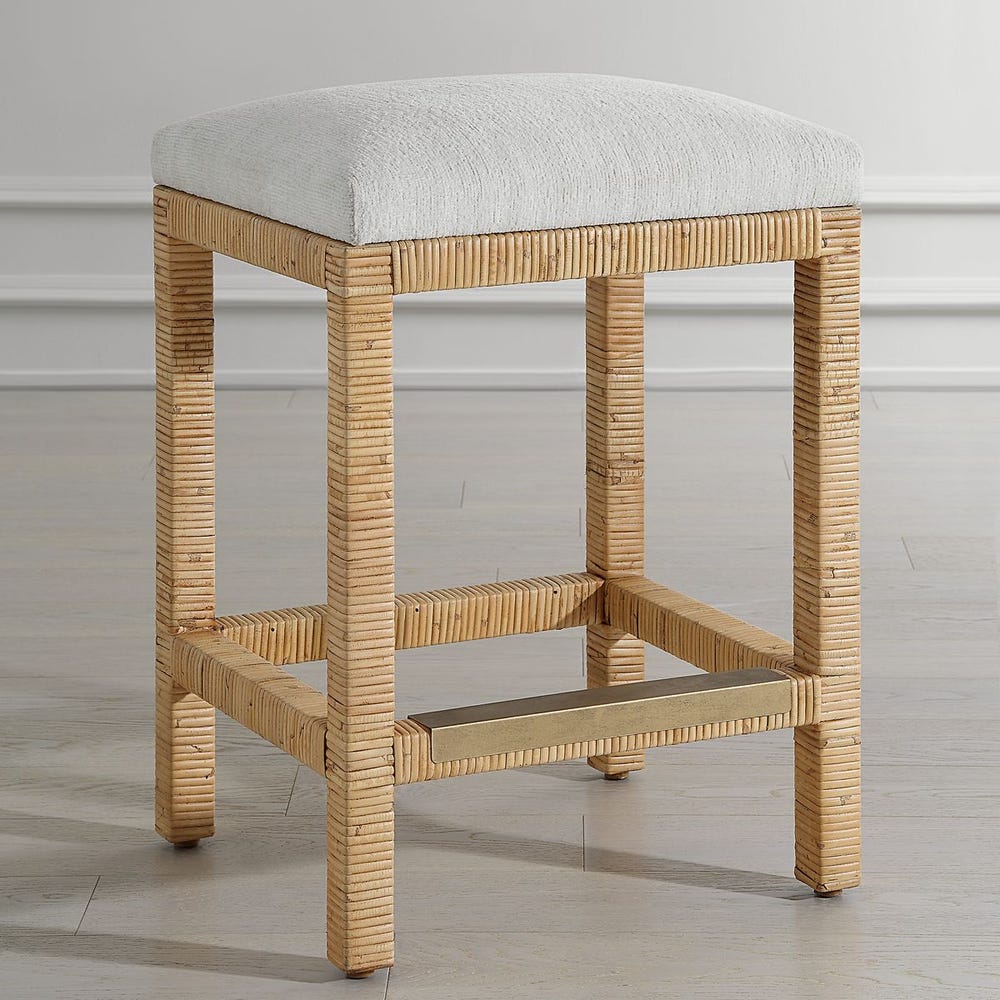 Handwoven rattan counter-height stool with off-white cushion and antique gold kickplate—coastal-inspired and refined.